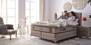 Noptieră modernă Ferremo LUXIA GLASS, tapițată cu material textil, cu blat superior din sticlă securizată și picioare înalte din lemn.